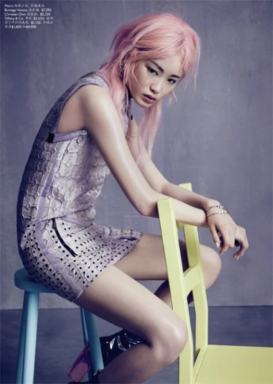 Young woman with pink hair wearing a purple dress sitting on a blue and yellow stool in a studio with a neutral gray background.