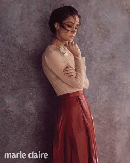Woman wearing sheer beige top and red leather skirt leaning against textured wall by Koh