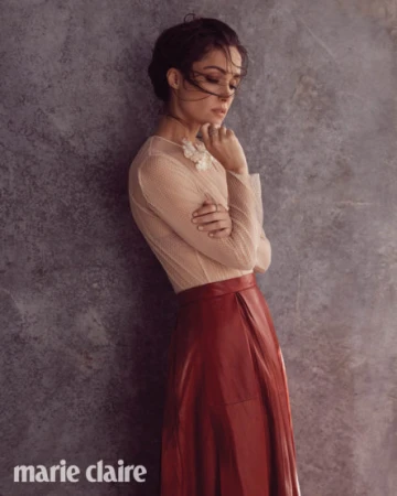 Woman wearing sheer beige top and red leather skirt leaning against textured wall by Koh