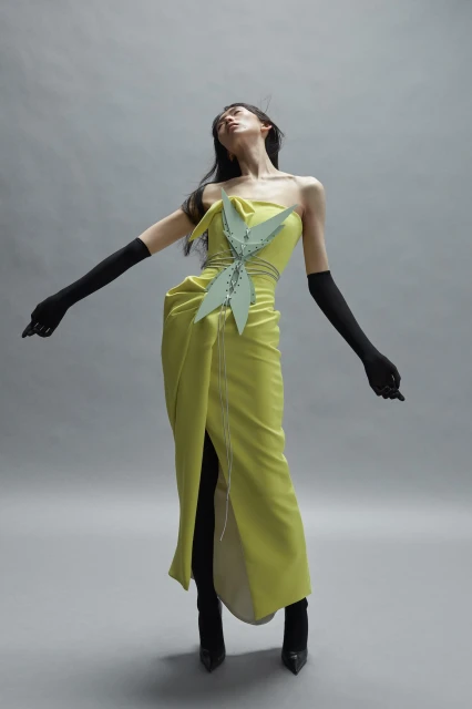 A full-length studio shot presents an Asian woman in a light-yellow, draped gown with black opera gloves, gazing upward in a light-grey studio.