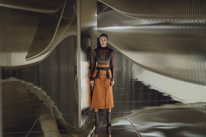 Full-length shot of a woman posing indoors with metallic, wavy walls in a fashionable, long-sleeved top and an orange skirt with knee-high boots.