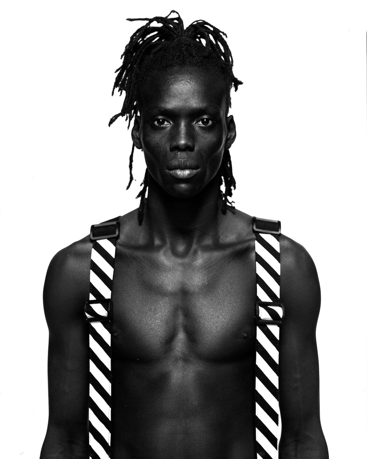 Striking black and white portrait of a shirtless Black male with dreadlocks wearing striped suspenders against a white backdrop.