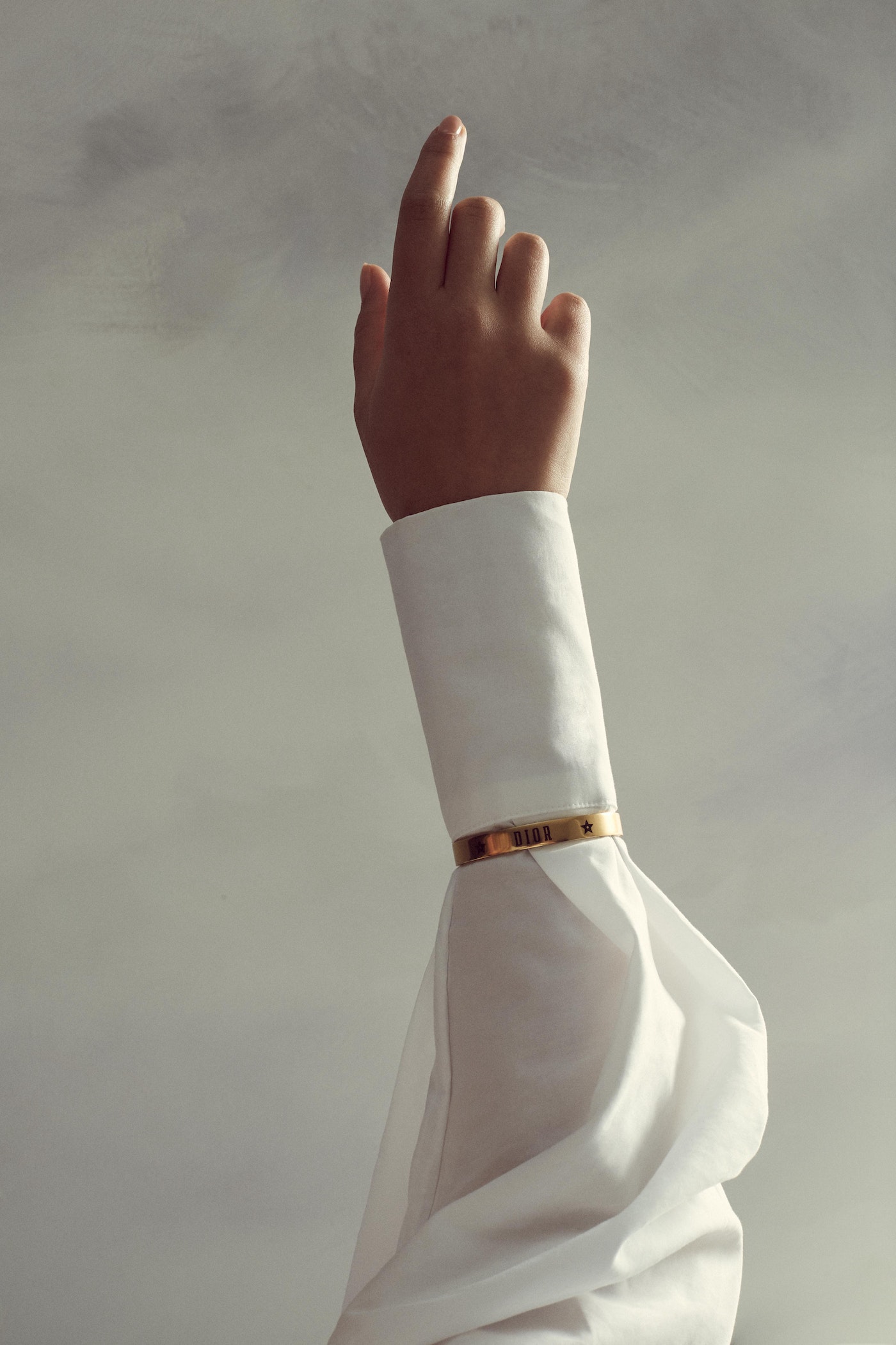 Elegant arm and hand in a white sleeve pointing upward against a soft grey background, accessorized with a delicate gold bracelet.