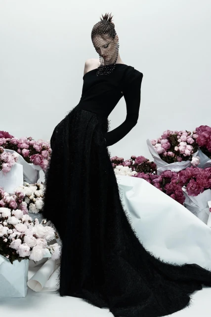 Elegant woman in a black, one-shoulder gown with a beaded headpiece, posing amid bouquets of pink flowers against a white backdrop.