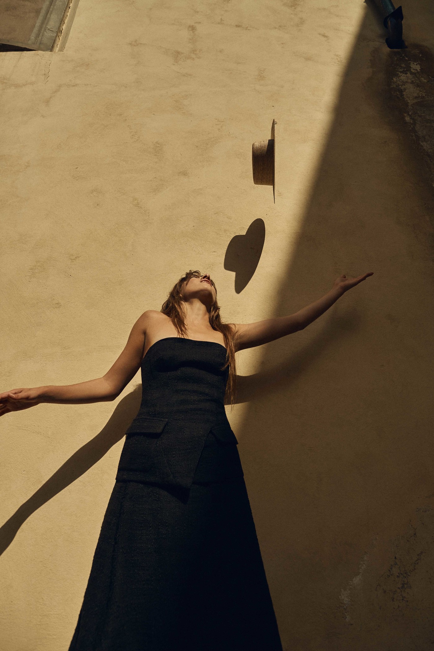 Young woman in black strapless dress reaches to catch a hat floating above in outdoor setting with textured yellow wall backdrop.