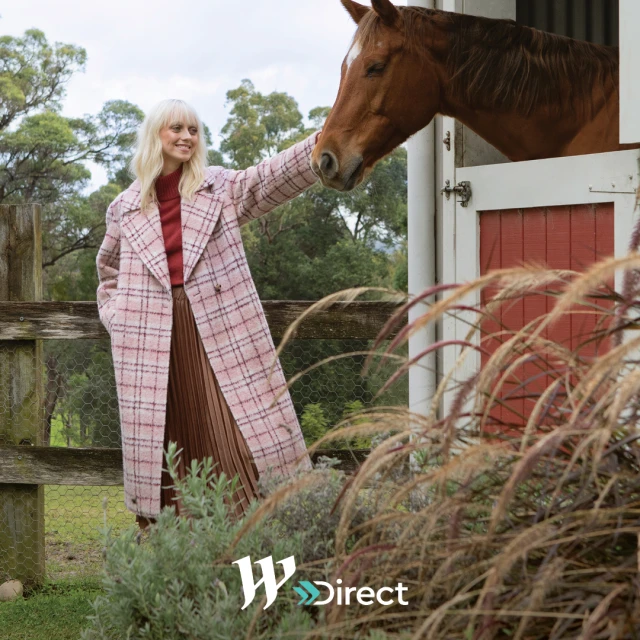 A woman in a pink coat stands next to a horse that is sticking its head out of a stable window in a rural setting.