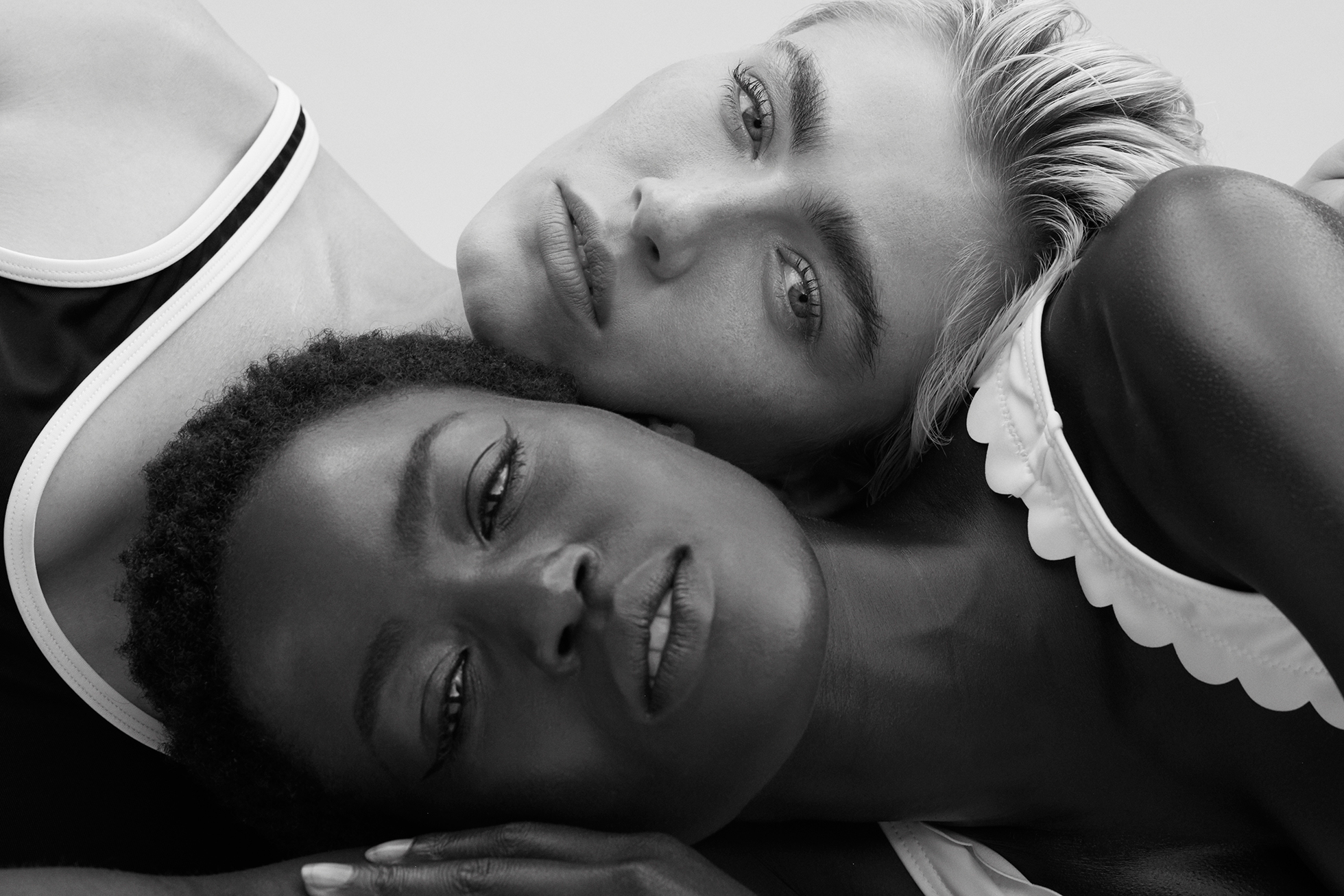 Black and white portrait of two diverse adult women posing closely together in swimwear against a neutral background in a photo studio.