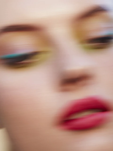 Close-up portrait of a young adult female face with vibrant makeup. Focus on pink lips, blue eye makeup, and a blurred effect.
