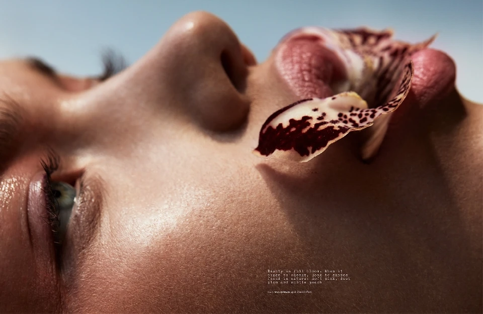 Close-up of a woman's face with an orchid petal on her lip, soft lighting, and a clear blue sky background. Beauty and skincare theme.
