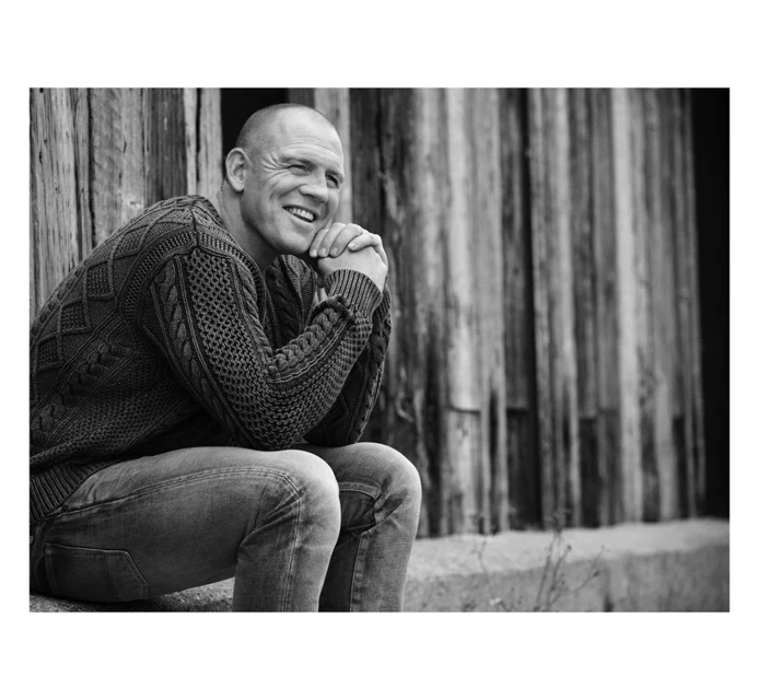 Black and white portrait of an adult male with short hair, wearing a knitted sweater, sitting outdoors against a rustic wooden background.