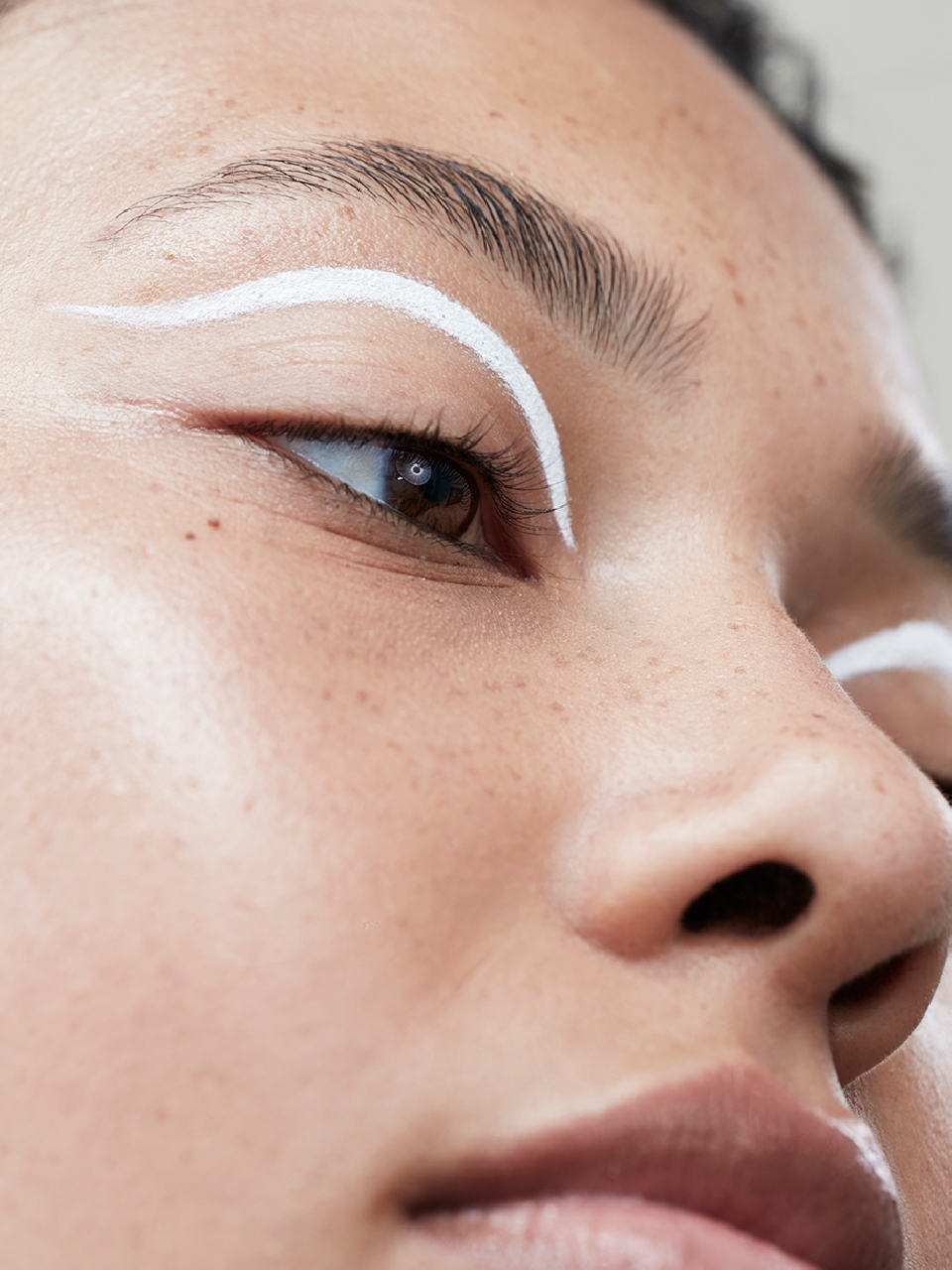 Close-up studio shot of a young adult woman's face with fair skin, light brown eyes, and white graphic eyeliner.