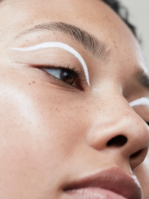 Close-up studio shot of a young adult woman's face with fair skin, light brown eyes, and white graphic eyeliner.