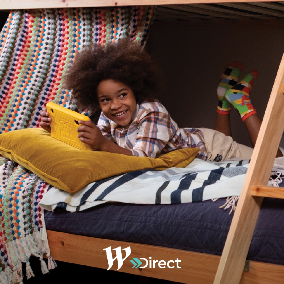 Child with afro laying on a bed underneath a wooden frame, smiling while using a yellow tablet, colorful blanket and pillow nearby.