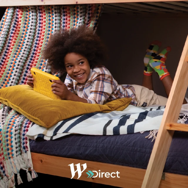 Child with afro laying on a bed underneath a wooden frame, smiling while using a yellow tablet, colorful blanket and pillow nearby.