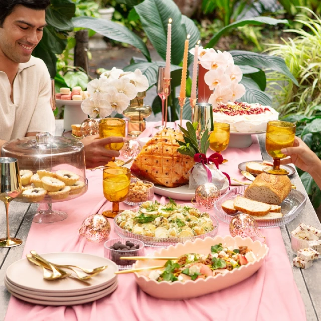 Outdoor dining table laden with food, pink tablecloth, surrounded by lush greenery, featuring a smiling man and amber glassware.