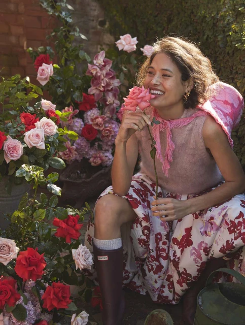 Young woman in a garden smiling, holding a pink rose, wearing a floral print skirt, Hunter boots, and a pink top on a sunny day.