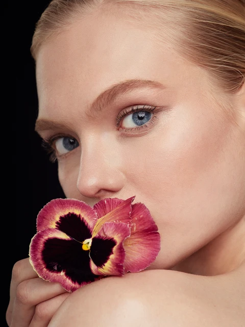 Close-up portrait of fair-skinned adult woman with blue eyes and blonde hair holding a pansy flower to her mouth, set against black background.