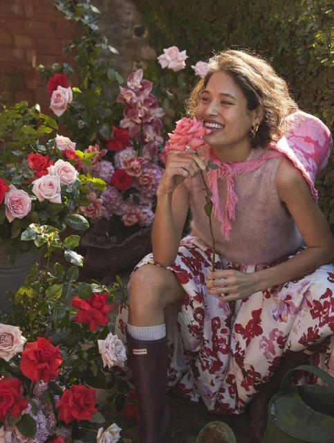 Woman smiles smelling a pink rose, seated among roses and flowers in a garden setting wearing floral skirt and rain boots.