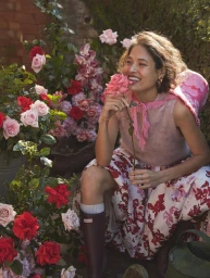 Smiling woman in a pink sleeveless top and floral skirt holding a rose in a garden by Jen Wilding