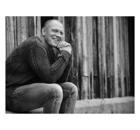 Black and white portrait of a smiling bald man seated against a weathered wooden wall, wearing a textured sweater and jeans.