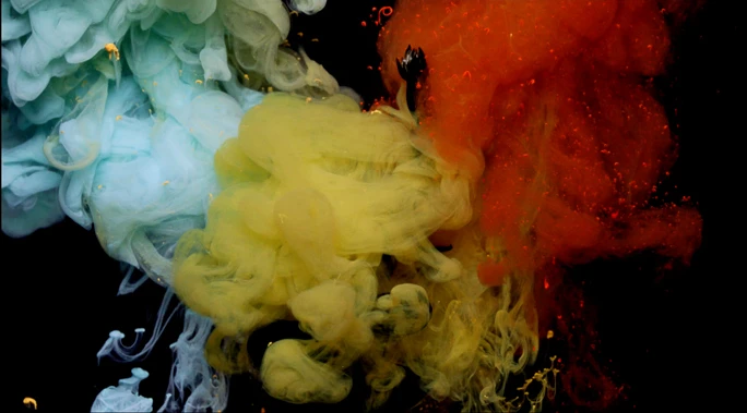 Abstract view of plumes of orange, yellow, and blue ink swirling and mixing in water against a stark black background.