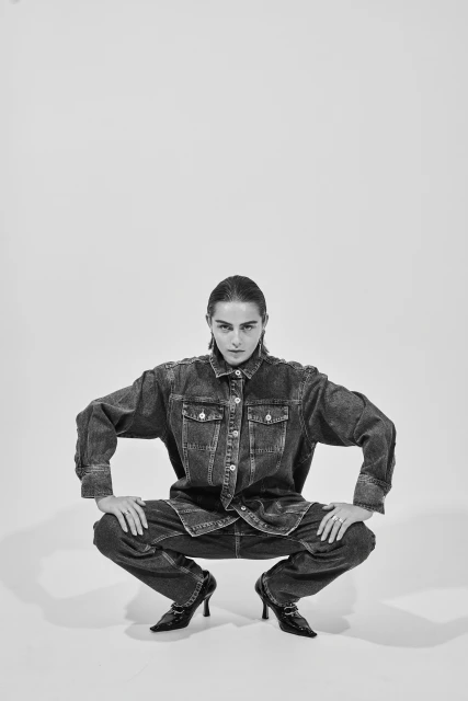 Woman poses crouching in studio wearing oversized denim jacket and jeans with heeled shoes, shot in black and white.