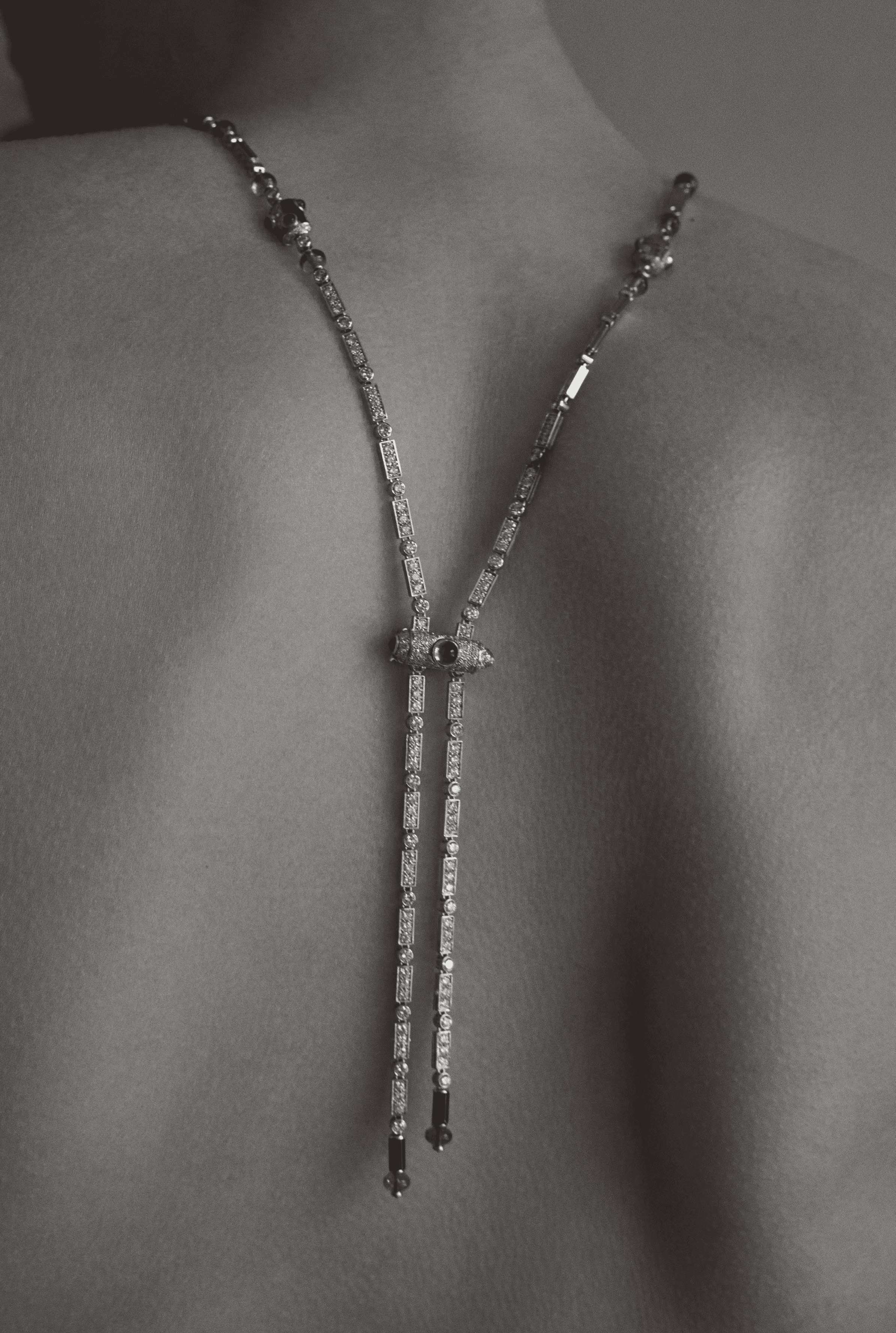 Close-up shot of a bare back adorned with a long, silver-tone necklace featuring geometric details, captured in soft, diffused lighting.