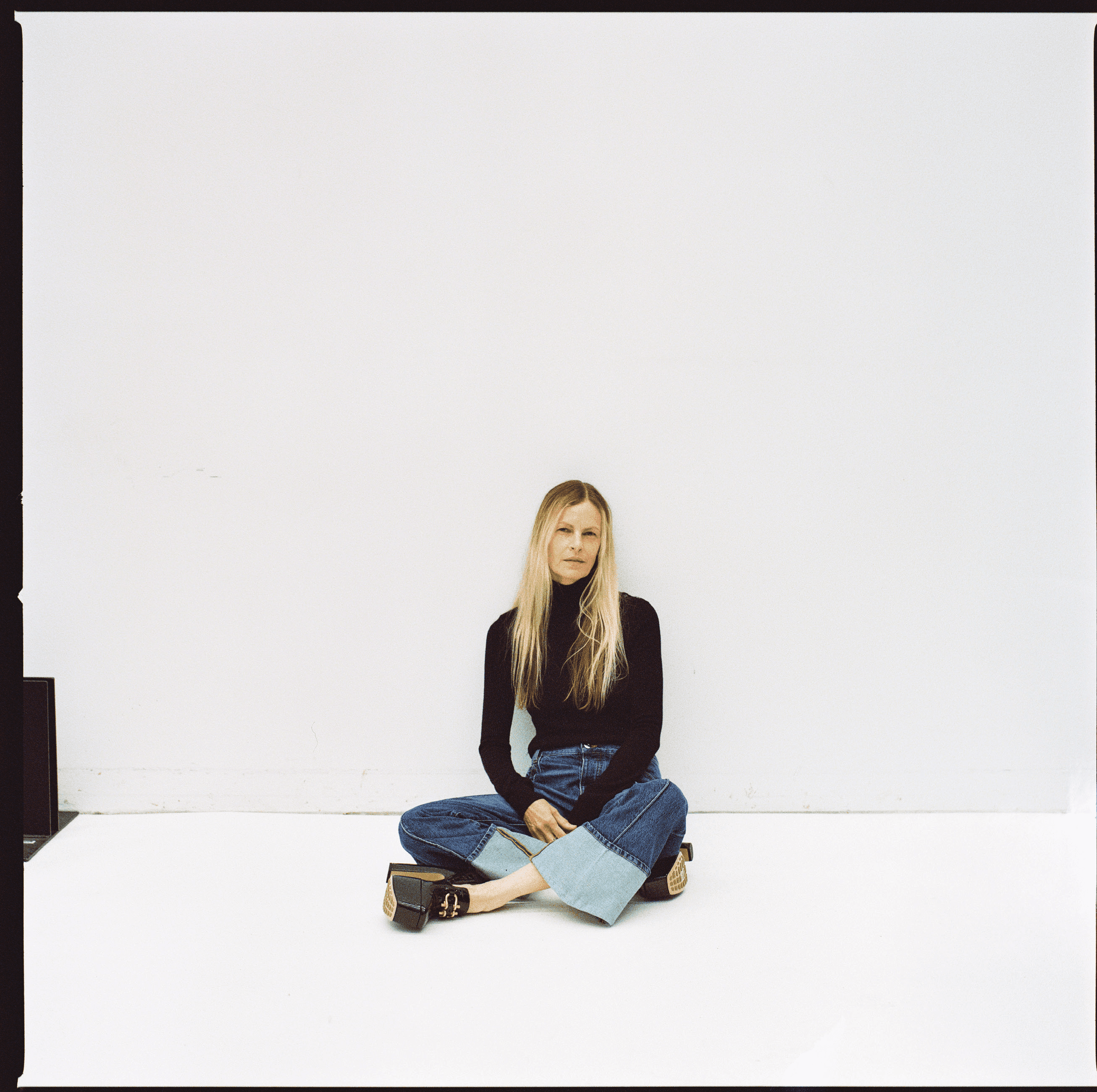 A young woman with long blonde hair sits cross-legged on a white surface wearing a black turtleneck and wide-leg denim jeans.