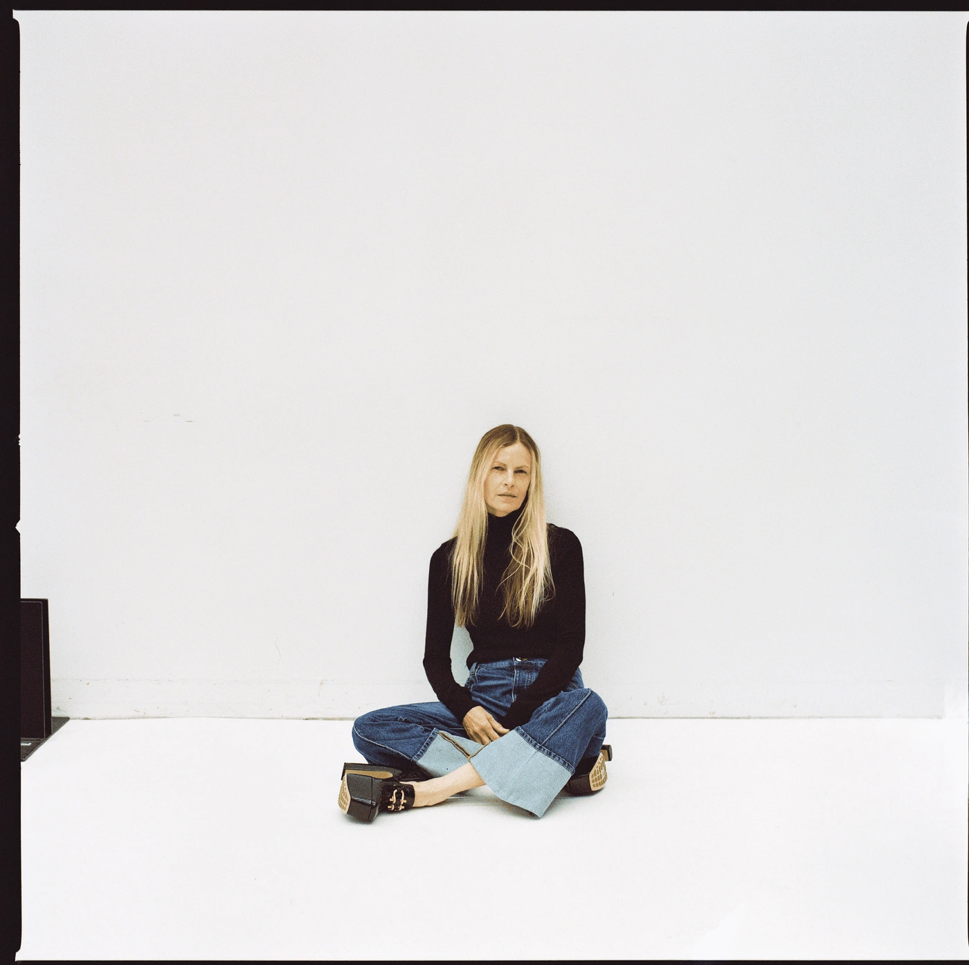 Woman with long blonde hair wearing a black turtleneck and cuffed blue jeans sitting on a white studio floor