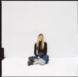 Woman with long blonde hair wearing a black turtleneck and cuffed blue jeans sitting on a white studio floor