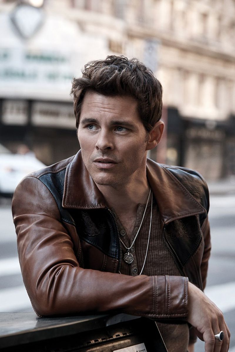 A man with brown hair poses in a brown leather jacket on a city street looking to the left with blurred buildings in the background.