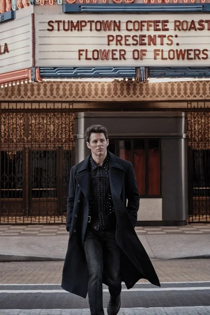 An adult male walks in front of a theater marquee. The man wears a black overcoat and black denim jeans on a city street.