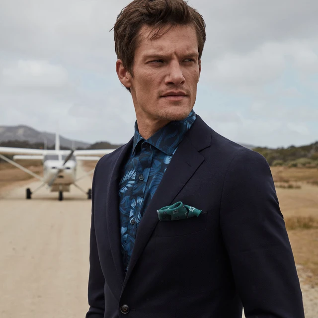 Stylish man in a navy suit and patterned shirt stands on a runway near a small airplane, looking off into the distance on a cloudy day.
