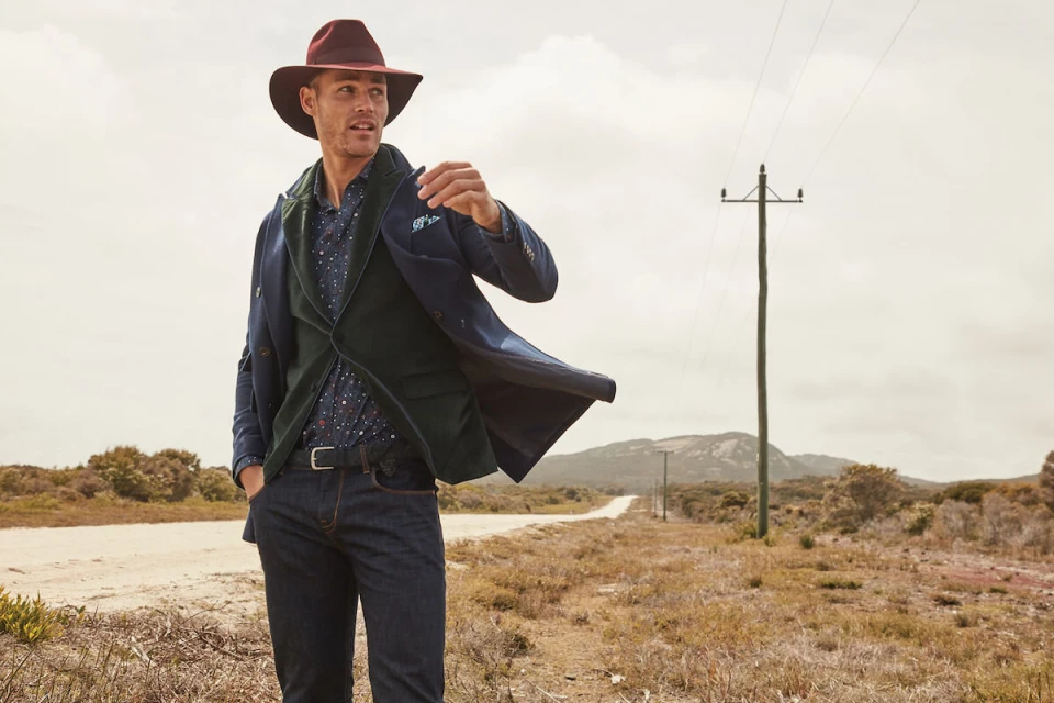 A male model in a stylish blazer, vest, button-down shirt, jeans, and wide-brimmed hat poses on a dirt road with telephone poles.