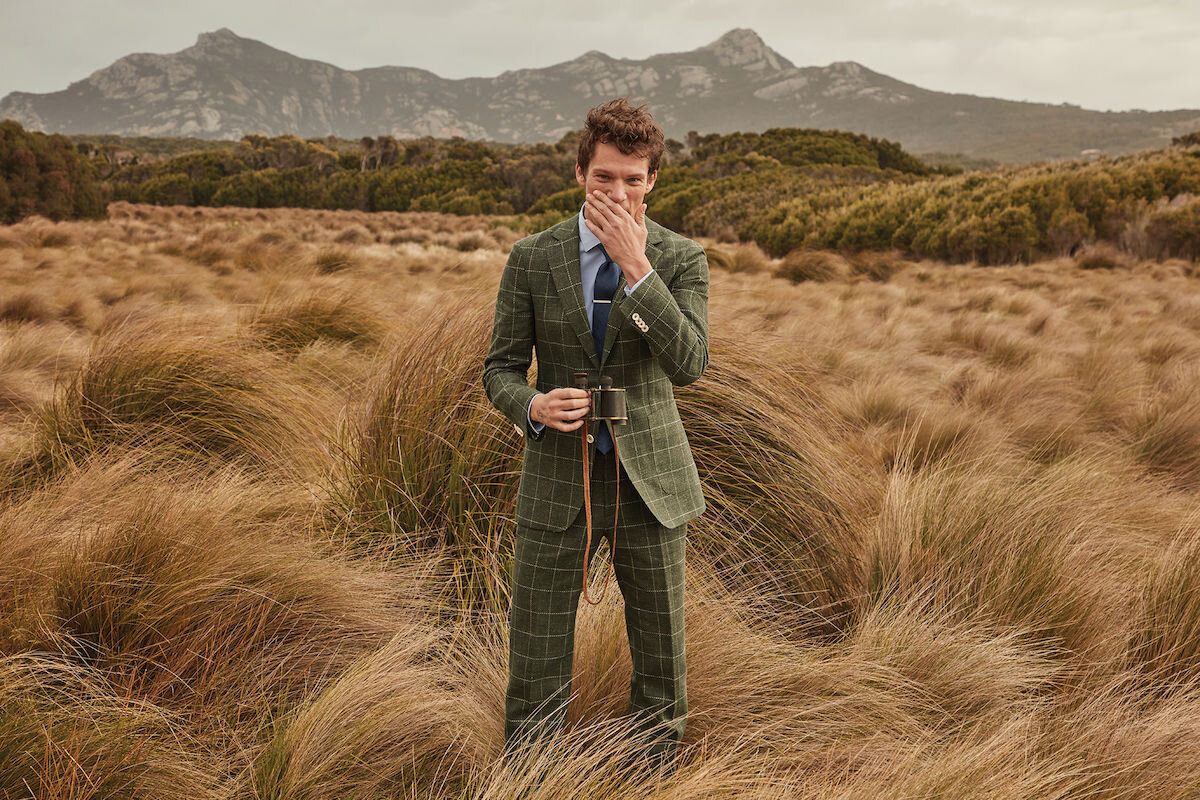 A man in a green suit stands in a grassy field holding binoculars, with mountains visible in the background beneath an overcast sky.