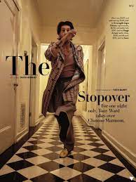 Fashion editorial photo: Adult male in patterned coat and trousers walking down a tiled hallway. Shot with eye-level angle and warm lighting.