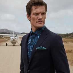 Man wearing a dark navy blazer and blue tropical print shirt with a small airplane parked on a dirt runway in the background.