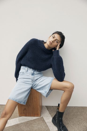 A woman in a navy sweater and denim shorts poses against a plain background, sitting on a wooden cube with black boots.