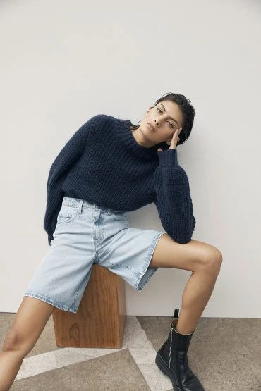 A woman in a navy sweater and denim shorts poses against a plain background, sitting on a wooden cube with black boots.