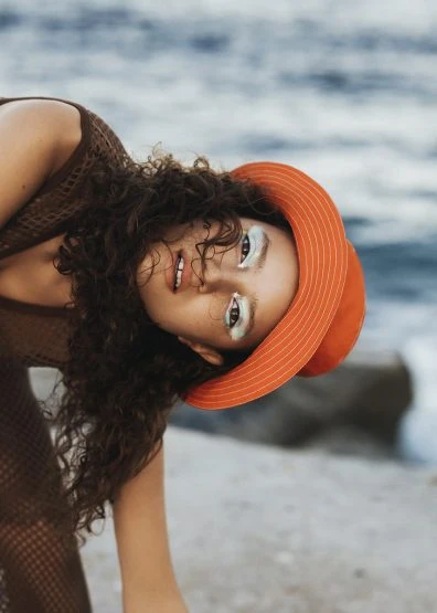 Portrait of a woman with curly hair and an orange hat posing on the beach with a natural, sunlit ambiance and neutral-toned clothing.