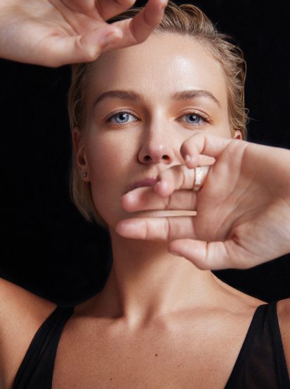 Close-up portrait of a blonde woman with blue eyes holding her hands up near her face against a black background.
