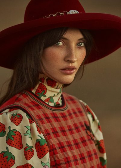 Portrait of woman with wide-brimmed hat. She wears a strawberry-print blouse with a plaid vest, shot in soft, diffused lighting.