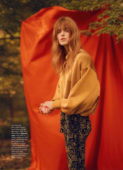A red-haired woman in a gold sweater and patterned pants stands outdoors in a fashion editorial shot against an orange backdrop.