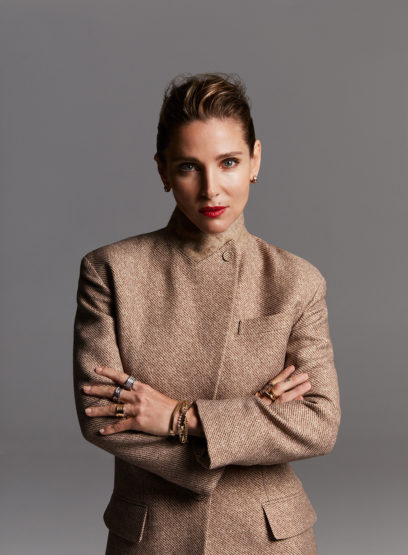 Portrait of a fair-skinned adult woman in a beige blazer, hands crossed, red lipstick, against a gray background. Fashion shoot.
