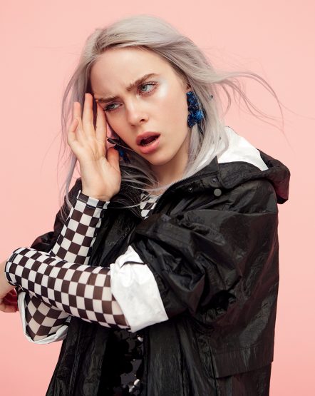 Close-up portrait of a young adult female model with gray hair and blue earrings wearing a black hooded jacket against a pink background.