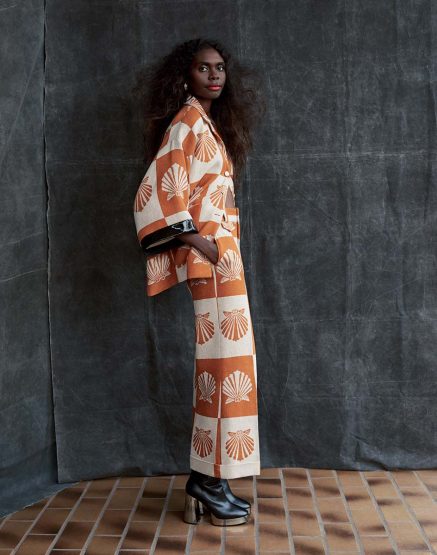 Woman poses in an orange and white seashell-patterned suit against a mottled dark gray background and warm-toned tiled floor indoors.