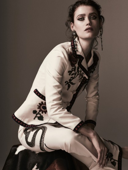 A woman with curled auburn hair wearing a cream-colored suit embroidered with flowers and snakes poses against a grey background.