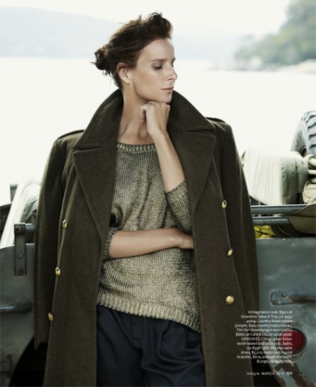 A brunette woman in an olive coat and gold sweater poses against an olive vehicle with a body of water in the background.