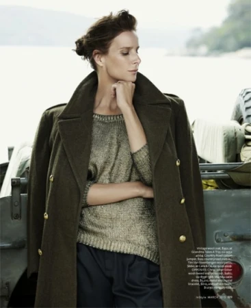 Model in olive green wool coat and metallic knit sweater standing against a vehicle outdoors with water background.