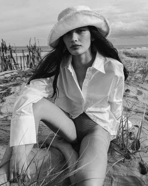 A woman in a white shirt and hat sits on a sandy beach with grasses in a black and white portrait, with a cloudy sky.